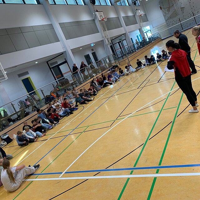 Schülerinnen und Schüler sitzen in der Sporthalle und hören die Erklärung zum Sportangebot