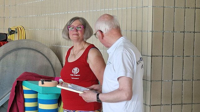 Zwei ältere Personen im Gespräch am Schwimmbadrand, eine Frau im roten Shirt mit Logo, ein Mann hält ein Klemmbrett.