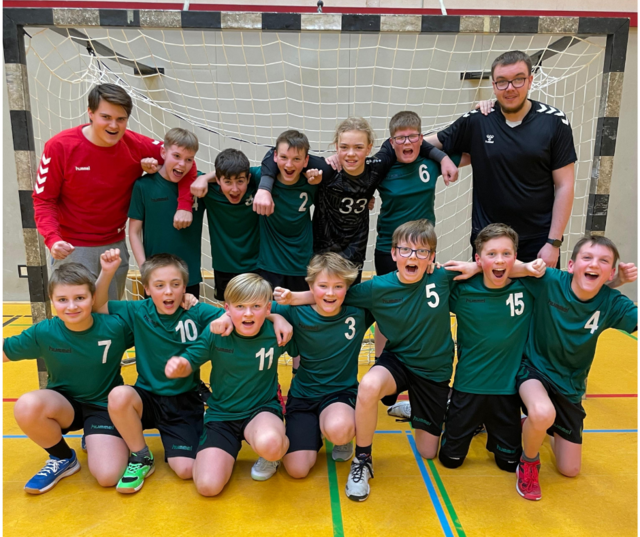 12 Jungen in grünen Trikots jubeln mit zwei Trainern vor einem Handballtor in einer Sporthalle.