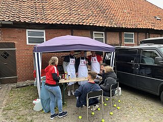 Drei Männer mit MTV-Schürzen stehen unter Pavillon am Grill, Gäste sitzen davor auf Stühlen mit Tennisbällen an den Stuhlbeinen.