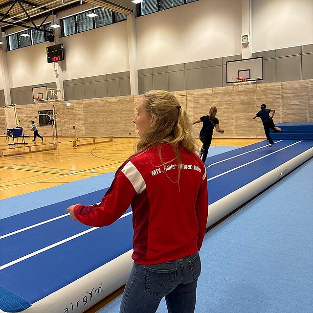 Betreuerin des MTV „Fichte“ erklärt in der Sporthalle das Airtrack, im Hintergrund laufen Schüler darüber