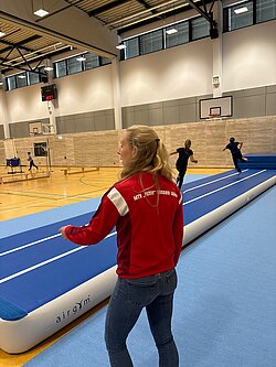 Betreuerin des MTV „Fichte“ erklärt in der Sporthalle das Airtrack, im Hintergrund laufen Schüler darüber