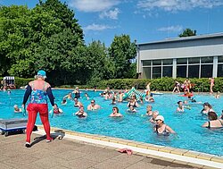 Große Gruppe bei der Wassergymnastik im Freibad, am Beckenrand leitet eine Übungsleiterin die Übungen an
