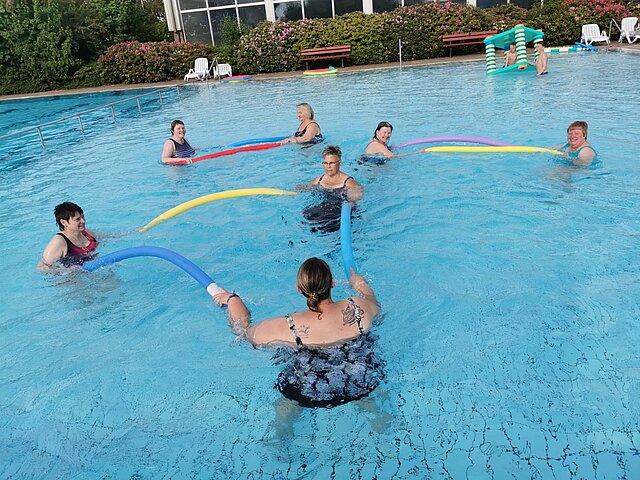 Rehasport im Wasser mit Poolnudeln Eine Übungsleiterin und sechs Teilnehmerinnen stehen im Wasser des Außenbeckens und halten Poolnudeln beim Rehasport