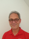 Älterer Mann mit grauen Haaren, Brille und rotem Poloshirt lächelt vor heller Wand in die Kamera.