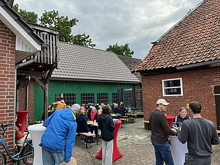 Menschen sitzen an Bierzeltgarnituren im Hof zwischen Backsteinhäusern und tauschen sich bei Snacks und Getränken aus.